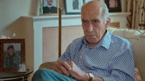 Mike Jackson sitting in an armchair with his legs crossed and hands sitting on his knee.
He is wearing a blue and white checked short and has grey hair.