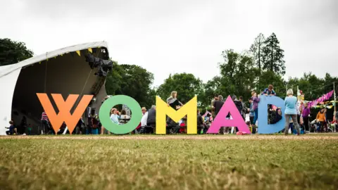 Getty Images A large, multi-coloured sign saying "Womad" is placed on grass. Behind is a small, covered stage for acts. People are milling around the sign.