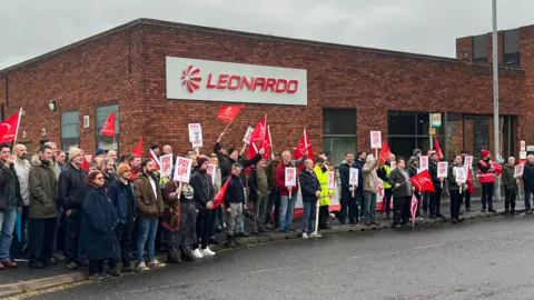 BBC A row of workers outside the exterior of Leonardo in Yeovil. They are waving red and white flags and placards. The Leonardo sign is on the wall above them.