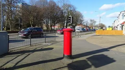 A line of cars approaching the crossing, which has railings on one side. A red post box is in the foreground, with a number of houses in the distance.