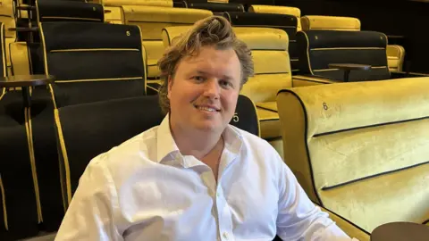 James Jervis sat in a seat in a cinema with black and gold chairs velvet chairs. He is wearing a white shirt and has thick brown hair and is smiling at the camera. 