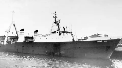 A black and white image showing a large trawler. The vessel is berthed. The upper deck is white, with the hull painted in a dark colour. 
