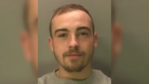 A man with a short beard and moustache and thin hair in a police mugshot.