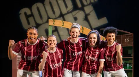 Five women wearing red and white football kits are smiling open-mouthed towards the camera and three are holding up fists in triumph