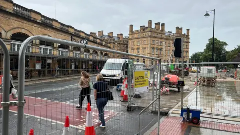 This is a picture of the ongoing work on York's Station Gateway scheme. Metal fences have closed off part of the area opposite the station entrance where new paving stones are being put in. People using the temporary pedestrian crossing are walking past traffic cones.