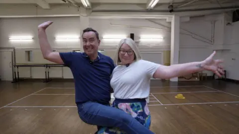 Edward Sault and Sophie Law are dancing with their arms outstretched and leg over one another.