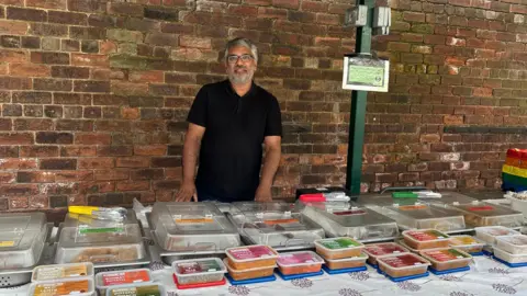 Jessica Bayley/BBC Stallholder Shan Ajaib at Pudsey Market stood behind a table with food products in brightly coloured boxes on his stall. He is wearing a black t-shirt with white writing on it and he has a beard.