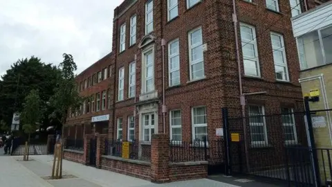 A large brick school building of at least three storeys has a sign saying "St Anne's" and is surrounded by railings