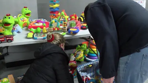 A man and a woman look at toys on a table. There are several green dinosaur toys. They are both wearing black tops.