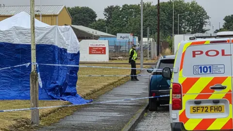 A police tent on a grassy area on an industrial estate. A a police cordon is in place at the front of the scene
