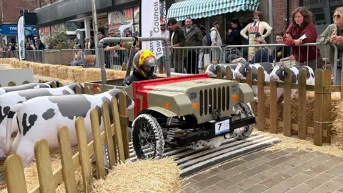 Barrow BID A soapbox derby driver crosses a recreation of a cattle grid on a racing course through Barrow town centre. Either side of the track there are straw bales and inflatable cows.