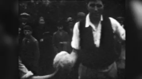 Unknown Black and white frame from a film showing a man in a white shirt and dark waistcoat holding a cabbage in his right hand. 