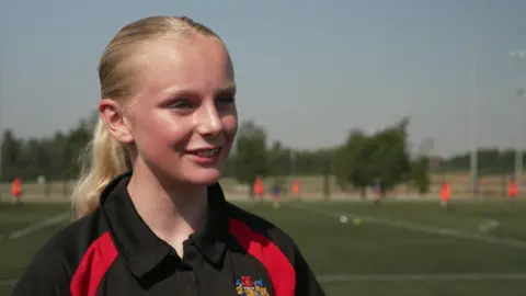 A girl of around 12, with blonde hair tied back into a pony tail and wearing a black polo-neck PE top, smiles at the camera in front of a football pitch