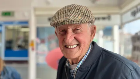 George Carden/BBC Dick Dixon smiling at the camera with the arcade behind him wearing a bomber jacket, flat cap and chequered shirt.