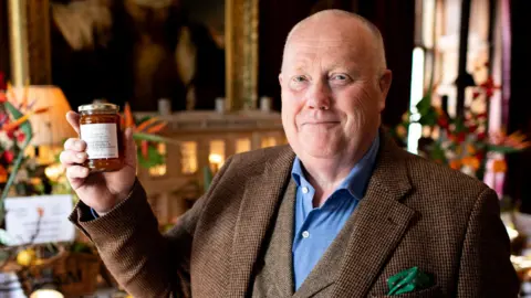 Hermione Mccosh David Hinkins is looking at the camera, smiling slightly, and holding his jar of marmalade aloft. He has short, fair hair and is wearing a brown suit. His shirt is blue and he has a green handkerchief in his top pocket. The jar has a white label and contains a dark orange marmalade.