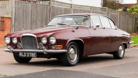 A maroon Jaguar car parked on tarmac with a wooden fence behind it