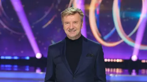 Getty Images A man with short, sandy hair wears a dark suit and polo neck shirt on the set of a TV show, he is smiling.