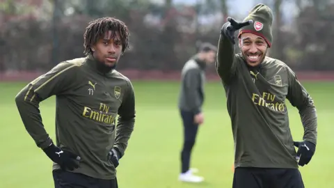 Alex Iwobi and Pierre-Emerick Aubameyang stand together on a green grass Arsenal training pitch, with Aubameyang, who is wearing an Arsenal woolly hat, pointing and pulling a silly face