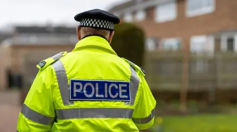 The back of a policeman wearing a hi-vis jacket standing in front of some houses. The back of his jacket says "police".