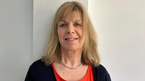 Amanda Ely Amanda has shoulder length blonde hair. She is wearing a dark cardigan and a red top with a gold necklace. She is smiling at the camera.