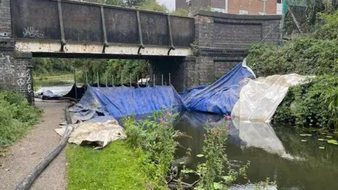 BBC Walsall Canal, with pipes and tarpaulin across it to assist clean up after a toxic spill, underneath a bridge at the top of the frame