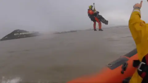 RNLI An RNLI crew member being winched above the water with the casualty strapped to him and the edge of a lifeboat in the foreground