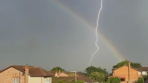 Lauren A view of a dark clouded sky above roofs of houses and the tops of trees. A downward lightning bolt crosses a faint but definite rainbow to the right of the image.