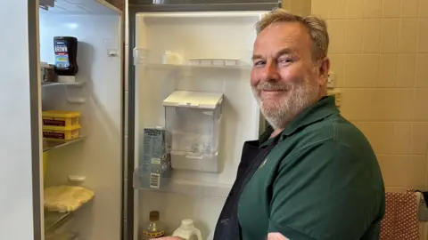 BBC/Lucy Ashton A man with short grey hair and a short grey beard is wearing a green polo shirt. He is smiling and standing by an open fridge door