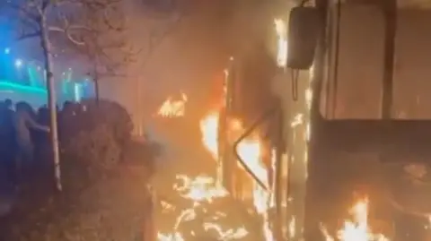 Vehicles burn on the right of the frame, as people are seen protesting on the left. It is night time