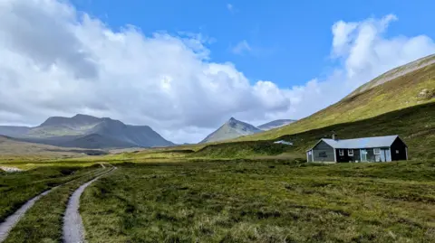 Alex Kintrea A long track winds along a grassy path. A building is on the right-hand side and two mountains are in the background
