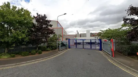 A view of a factory in an industrial estate taken from the road. Blue gates are across the entrance and a HGV is in the background.
