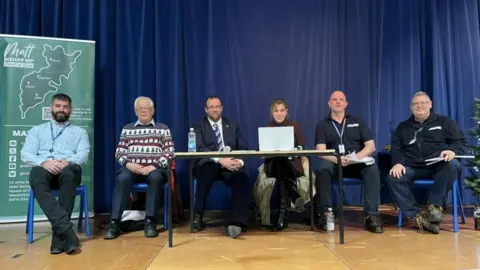 Six people are sitting on chairs on a stage - five men and one woman. In the middle is Matt Bishop, the Forest of Dean MP. To the left of him, in a Christmas jumper, is Roger Whyborn, Liberal Democrat councillor on Gloucestershire County Council. Taking notes on the right of Matt Bishop is his assistant. There is a blue velvet curtain behind the people on the stage as well as a map of the region.