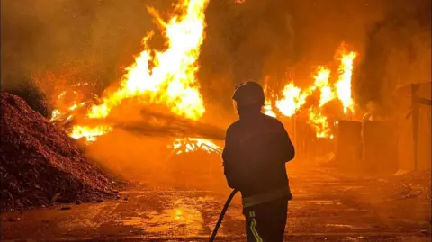 A picture of a Devon & Somerset Fire & Rescue Service firefighter tackling a large fire with bright orange flames in the dark. He has a water hose and is spraying it on the flames. 