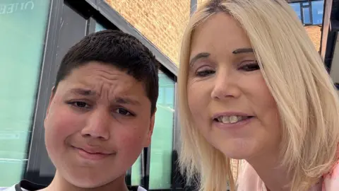 PA Media Reece Khan and his mother Selina Niman. Reece has short black hair and is looking at the camera. His mum is stood on the right of the photo and has shoulder-length blonde hair and is smiling at the camera. They are stood outside and buildings can be seen in the background.