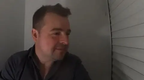 Richard Gill/BBC Kevin Shoesmith, a journalist in his mid 40s, with cropped brown hair, is wearing a grey open neck shirt. He is looking nervous, hiding from a dog in a small cupboard in a former police station.