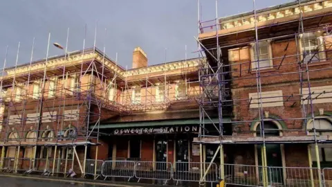 A three-storey brick building with arched windows and and canopy above the main entrance reading "Oswestry Station". The entire building is covered in scaffolding. 
