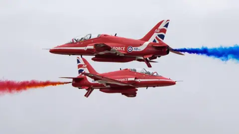 PA Media Two red Royal Air Force planes fly through the sky. The plane on the bottom shoots out red smoke to the left, while the plane on the top shoots out blue smoke to the right.