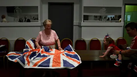 PA Media A woman is folding a union jack flag in a hall, a little girl being held up by an adult is attempting to crawl across the table to the flag. The girl is wearing a red top and trainers with blue socks. 