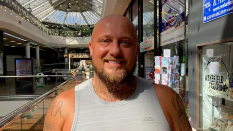 A bald man with a blond-brown beard and neck and arm tattoos wearing a grey vest 