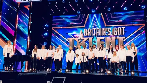 A large choir, featuring adults and children of all ages, on stage in front of the Britain's Got Talent logo.