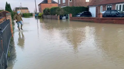 A flooded street which shows water covering the road and gardens of the surrounding properties. A man wearing green wellies, blue jeans and green jacket is wading through the water on the left of the image. He is looking at the house which is surrounded by water and has two black cars parked outside.