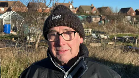 Oliver Conopo/BBC A man wearing a black winter hat and glasses. He is standing in an allotment with the backdrop of new build houses on a bright winter day.