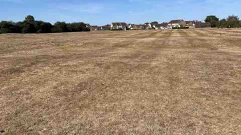 John Devine/BBC A large expanse of parched grass which undulates like the waves on a sea, a fort once stood on this land, houses can be seen in the far background.