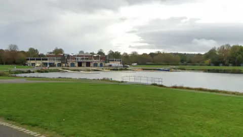 Google A lake with a large building at the end. It is surrounded by grass. There are four yellow floating piers on the water.