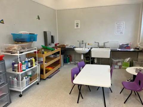 A mock-up of a nursery crafting area with paints on a trolley, a white table, four purple chairs and two hand-washing sinks. 