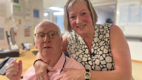 Photo of Norman and Christine taken in a hospital area but not a ward. Norman is sat down and Christine is standing. She is leaning over Norman with her hands around his shoulders. He is bald-headed and is wearing glasses with a yellow tint. He has a pink t-shirt. Christine has short blonde hair and is wearing a sleeveless patterned shirt. She is displaying a watch on her arm. 