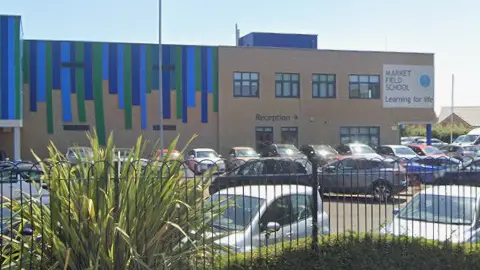 The Market Field School building is two levels high and mostly beige, although there are blue and green lines on part of it. Cars are parked in front of the building.