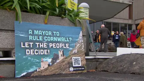 A sign reading "A Mayor to rule Cornwall - let the people decide" in front of protestors outside County Hall. 