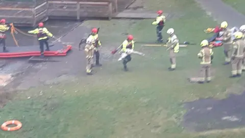 Guy Booth Fourteen firefighters (some out of shot) wearing fluorescent clothing attempt to rescue a swan from a frozen pond.