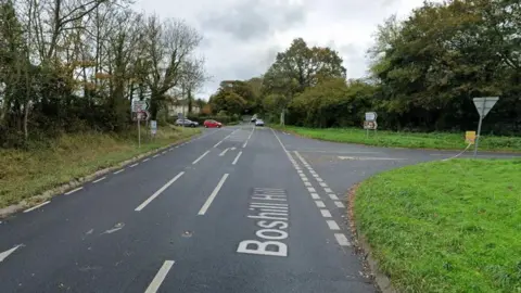 Google Street View image of Boshill Cross on the A3052 in Colyford. A couple of cars are driving along the road, facing the camera. Two cars are waiting at a T-junction on the left hand side.